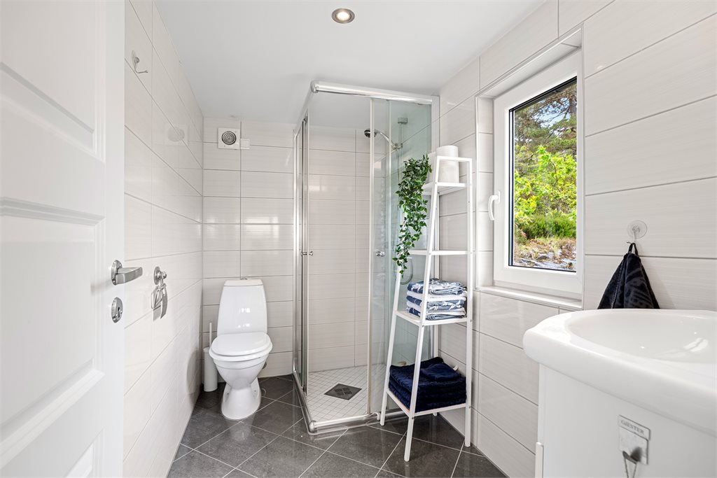 Badet fremstår lyst og moderne med flislagte overflater, dusjhjørne, servantinnredning og toalett.